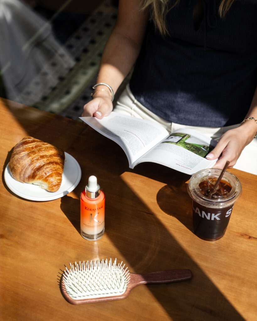 Woman enjoying breakfast and reading a book at home with the Kérastase Nutritive Scalp Serum and Fabio Scalia La Claudia brush on the table, representing a calm morning self-care and scalp wellness ritual.